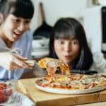 Crop delightful Asian ladies smiling while cutting piece of delicious homemade pizza with stretched cheese on cutting board in kitchen