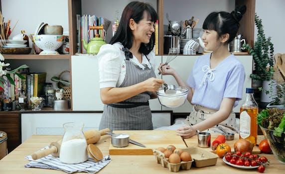 Happy Asian mother wearing apron and daughter standing near wooden counter with ingredients while discussing recipe of pizza