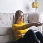 Joyful millennial female in casual clothes with bowl of snack using remote controller while sitting on floor leaning on sofa and watching movie in cozy light living room with luxury interior