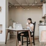 Pensive young barefoot female sitting on chair with bending leg and watching video on laptop while resting and drinking hot coffee at table in modern light apartment