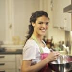 Side view of cheerful female in apron and casual t shirt standing in modern kitchen and mixing ingredients with whisk in stainless bowl while preparing dough