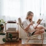 Cheerful young female in eyeglasses with cup of beverage reading textbook in armchair between potted plants in house room