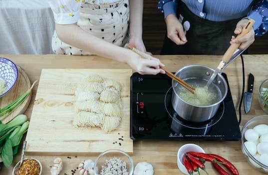 Crop anonymous housewives in aprons boiling noodles on tabletop stove near fresh ingredients in modern kitchen