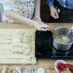 Crop anonymous housewives in aprons boiling noodles on tabletop stove near fresh ingredients in modern kitchen