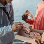 Crop anonymous male friends in casual wear eating delicious pizza together on embankment