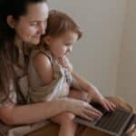 Crop side view of adult smiling mother embracing little daughter while typing on netbook and sitting in room in flat