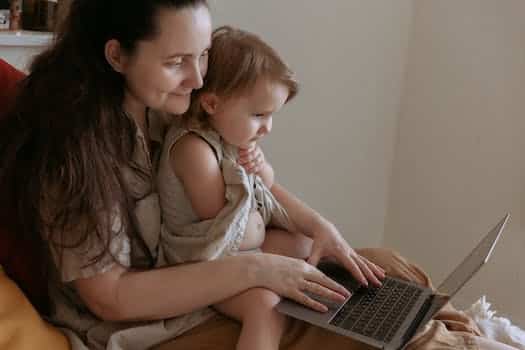 Crop side view of adult smiling mother embracing little daughter while typing on netbook and sitting in room in flat