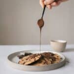 Crop unrecognizable chef pouring chocolate syrup on pancakes in plate on table on gray background