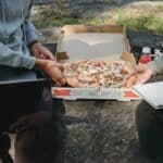 Faceless crop young men spending time on boulder outside with computer and pizza and with notepad in summer sunny day wearing casual clothes