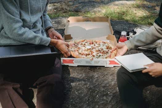 Faceless crop young men spending time on boulder outside with computer and pizza and with notepad in summer sunny day wearing casual clothes