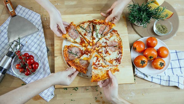 From above unrecognizable people taking slices of pizza with salami melted cheese and herbs from plate on kitchen table