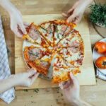 From above unrecognizable people taking slices of pizza with salami melted cheese and herbs from plate on kitchen table