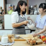 Happy Asian mother wearing apron and daughter standing near wooden counter with ingredients while discussing recipe of pizza