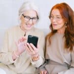 Happy elderly elegant trendy women looking at screen of mobile phone and shopping online while resting on sofa