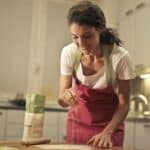 Low angle of positive young female in apron sprinkling flour over table while preparing food in modern light kitchen