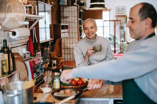 Male seasoning delicious meatballs in pan against cheerful female beloved with cup of coffee in kitchen