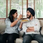 Positive Indian female in casual wear sitting on sofa in living room and feeding bearded male in turban