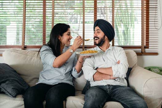 Positive Indian female in casual wear sitting on sofa in living room and feeding bearded male in turban