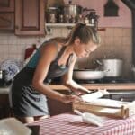 Side view of housewife wearing apron standing at table in cozy kitchen and preparing dough for baking while using rolling pin