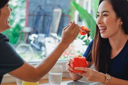 Side view of positive Asian female feeding smiling friend with salad while sitting with cold cocktail in restaurant near window with blurred street