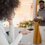 Smiling black man standing in room with bowl of salad near girlfriend sitting on couch