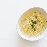 Top view of white bowl with yummy homemade noodles cooked in fresh delicious chicken broth and topped with green aromatic parsley placed on white marble table