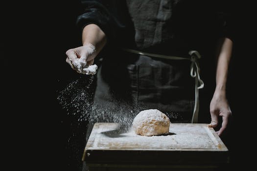 Unrecognizable baker in uniform standing at table and sprinkling flour in dough while cooking against black background