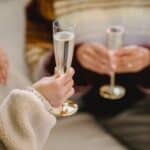 Unrecognizable women in casual clothes celebrating holiday sitting on sofa with glasses of champagne