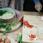 From above anonymous females cutting boiled eggs on wooden cutting board while cooking traditional Asian noodle soup in kitchen