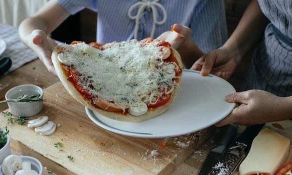 High angle of crop anonymous women transferring uncooked yummy homemade pizza on platter during preparation to baking in kitchen