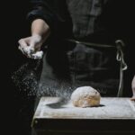 Unrecognizable baker in uniform standing at table and sprinkling flour in dough while cooking against black background